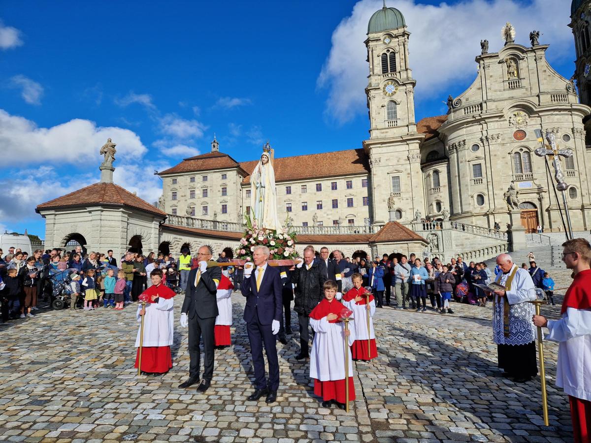 Männer in dunklen Anzügen tragen eine blumengeschmückte Marienstatue über einen gepflasterten Platz vor einer barocken Klosterkirche, daneben Ministranten in roten Gewändern und ein Priester mit Stola.